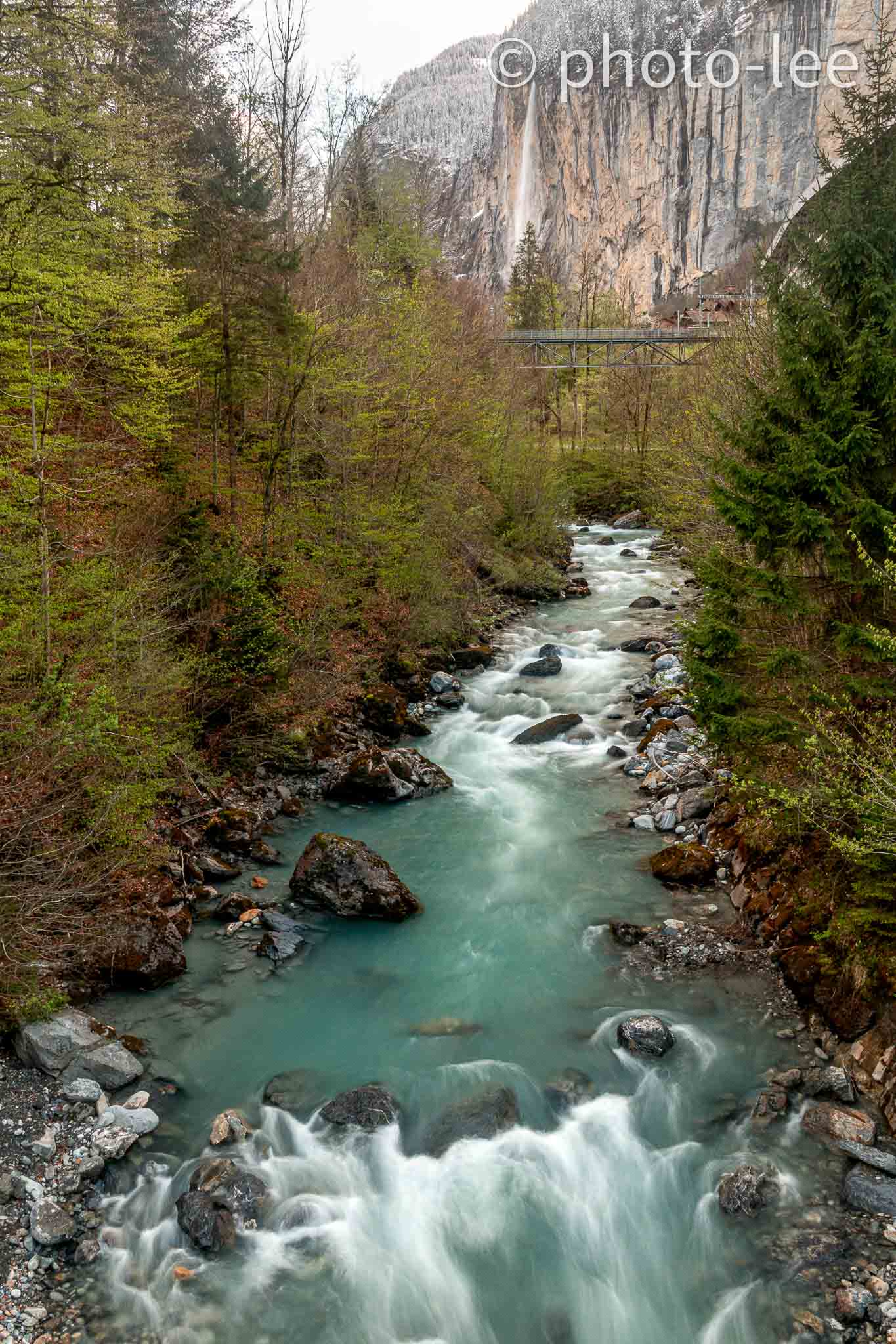 Vor einem langen Wasserfall fließt ein reißender Fluss zwischen halbgrünen Bäumen